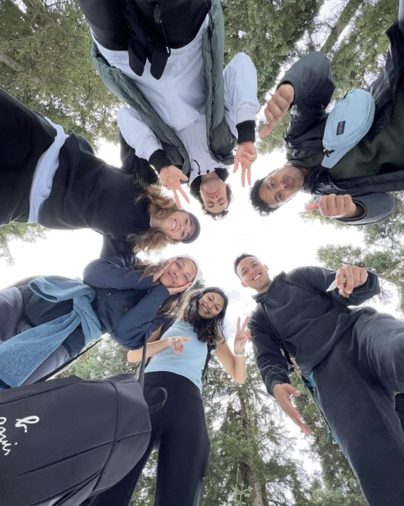 A group of students stand in a circle, smiling down at the camera, with trees in the background